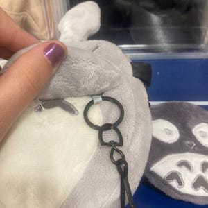 Op de afbeelding: Een pluche grijze en witte Totoro-vormige sleutelhanger. De sleutelhanger heeft een zwarte metalen ring en sluiting. Het pluche speelgoed heeft geborduurde gezichtskenmerken en een klein oor. Een ander soortgelijk pluche speelgoed is zichtbaar op de achtergrond.