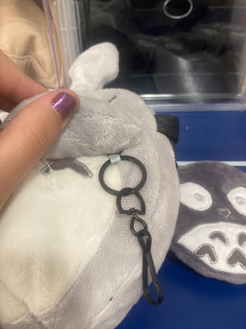 Op de afbeelding: Een pluche grijze en witte Totoro-vormige sleutelhanger. De sleutelhanger heeft een zwarte metalen ring en sluiting. Het pluche speelgoed heeft geborduurde gezichtskenmerken en een klein oor. Een ander soortgelijk pluche speelgoed is zichtbaar op de achtergrond.