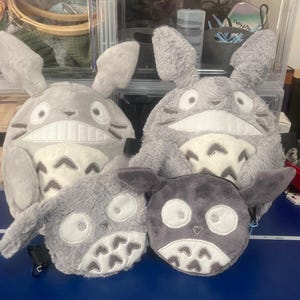 Op de afbeelding: Een verzameling grijze pluche tassen en pouches, ge&iuml;nspireerd op Totoro. De tassen hebben grote oren, witte buiken en geborduurde gezichten. De pouches zijn kleiner, met vergelijkbare ontwerpen. De items worden gepresenteerd op een blauw oppervlak.