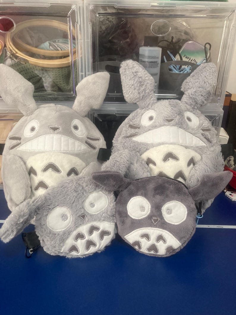 Op de afbeelding: Een verzameling grijze pluche tassen en pouches, ge&iuml;nspireerd op Totoro. De tassen hebben grote oren, witte buiken en geborduurde gezichten. De pouches zijn kleiner, met vergelijkbare ontwerpen. De items worden gepresenteerd op een blauw oppervlak.