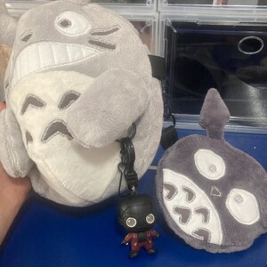 Op de afbeelding: Een grijze pluche Totoro-rugzak met een witte buik en een zwarte band. Een klein Deadpool-beeldje is met een zwarte clip aan de rugzak bevestigd. Er is ook een grijze pluche roet-sprite aanwezig.