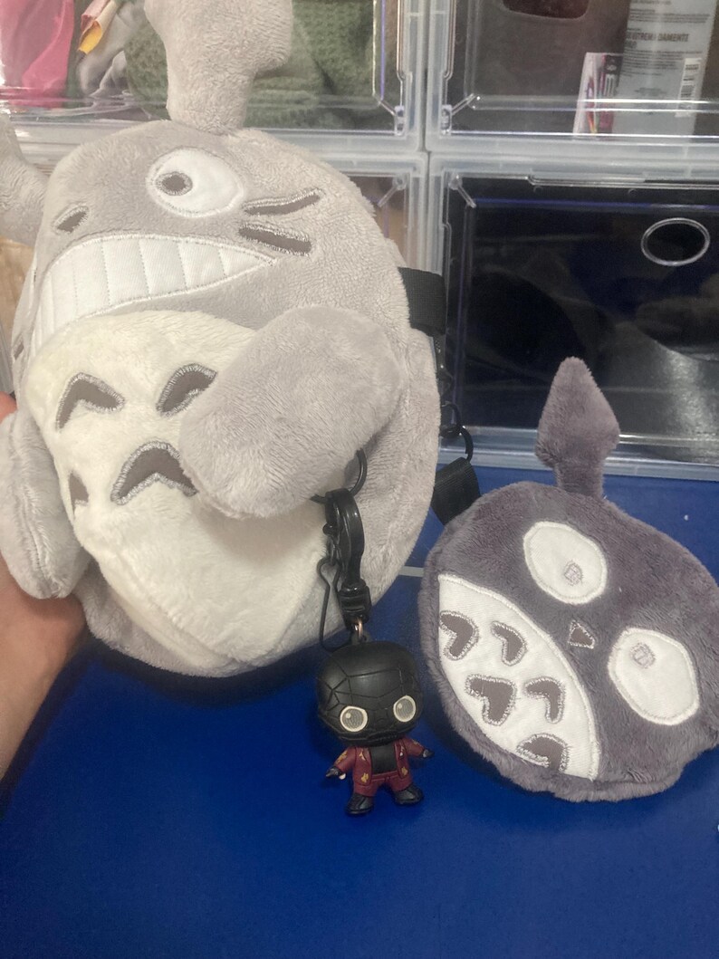 Op de afbeelding: Een grijze pluche Totoro-rugzak met een witte buik en een zwarte band. Een klein Deadpool-beeldje is met een zwarte clip aan de rugzak bevestigd. Er is ook een grijze pluche roet-sprite aanwezig.