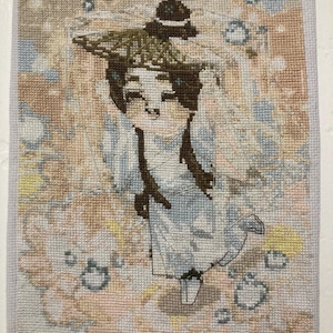 May include: A cross-stitch embroidery on white fabric depicting a figure wearing a conical hat and light blue robes. The figure is surrounded by soft, abstract patches of peach, yellow, and blue colors, with small circular shapes scattered throughout.