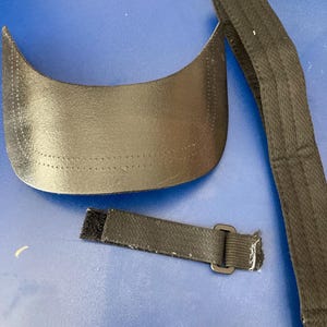 May include: A black visor with a curved brim and a matching adjustable strap. The visor is made of a smooth, possibly leather-like material. The strap has a buckle and a velcro closure.