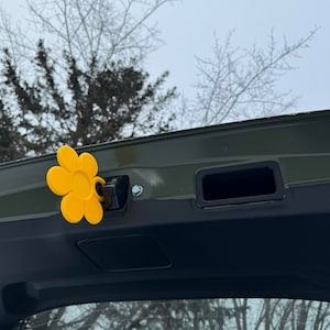 Puede incluir: Un amuleto de coche amarillo con forma de flor sujeto al cierre del maletero de un coche verde oscuro.