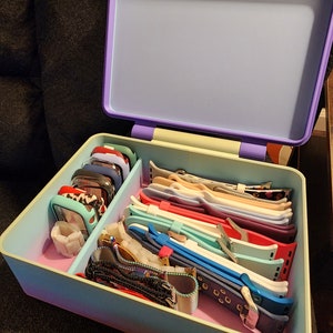 Puede incluir: Una caja de almacenamiento púrpura y amarilla con una variedad de correas y esferas de reloj en su interior. La caja está abierta y las correas están ordenadas de forma ordenada.