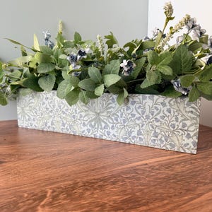 May include: A rectangular floral box with a light blue and green pattern, filled with green leaves and small blue and white flowers. The box sits on a wooden surface, creating a natural aesthetic.