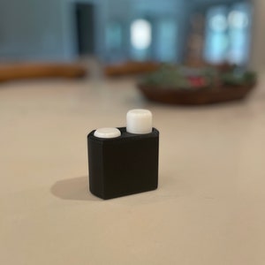 May include: A black plastic container with two white round lids. The container is on a white surface.