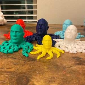 May include: A collection of colorful 3D-printed octopus figurines, each featuring a human face. The octopuses are in various colors, including green, red, blue, yellow, and white. The figurines are displayed on a wooden surface.