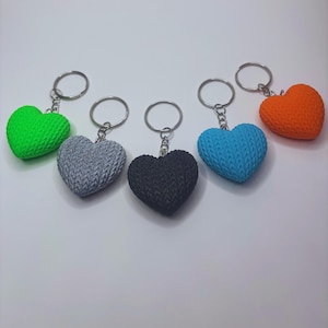 May include: A collection of heart-shaped keychains in various colors: green, gray, black, blue, and orange. Each keychain features a textured, knitted-like design and a silver keyring. The keychains are arranged on a white surface.