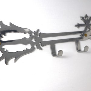 Puede incluir: Un gancho de pared de plata en forma de llave con un diseño decorativo. El gancho tiene dos puntas para colgar objetos.