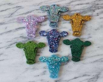 May include: Eight cow-shaped ornaments with glitter finishes in various colors, including blue, green, pink, and gold.