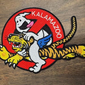 May include: A red and white circular patch with the text "KALAMAZOO" in white. A white ghost with a pink tongue is riding a yellow and orange tiger. The tiger has black stripes and is jumping.