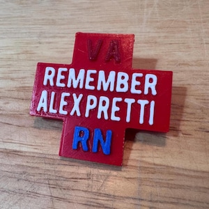 May include: A red cross-shaped badge with white text reading "REMEMBER ALEX PRETTI" and "VA" at the top. The letters "RN" are in blue at the bottom. The badge is on a wooden surface.