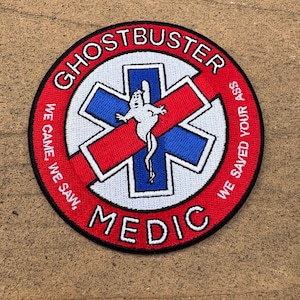 May include: A circular embroidered patch with a red border and background. It features a blue and white Star of Life symbol with a white ghost figure in the center. The text reads GHOSTBUSTER MEDIC and WE CAME, WE SAW, WE SAVED YOUR ASS.