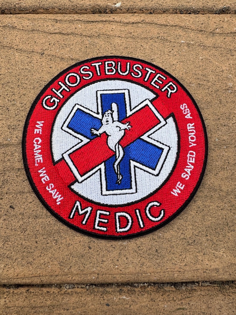 K&ouml;nnte beinhalten: Ein kreisf&ouml;rmiger gestickter Aufn&auml;her mit rotem Rand und Hintergrund. Er zeigt ein blau-wei&szlig;es Stern des Lebens-Symbol mit einer wei&szlig;en Geisterfigur in der Mitte. Der Text lautet GHOSTBUSTER MEDIC und WE CAME, WE SAW, WE SAVED YOUR ASS.