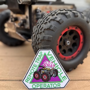 Könnte beinhalten: Ein dreieckiger Aufnäher mit dem Text "REMOTE TRAP VEHICLE OPERATOR" in Grün und Weiß. Er zeigt ein Cartoon-Fahrzeug mit lila Rädern und einer rosa Explosion. Der Aufnäher hat einen schwarzen Rand, vor einem verschwommenen Spielzeugfahrzeug-Hintergrund.