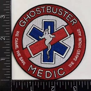 K&ouml;nnte beinhalten: Ein kreisf&ouml;rmiger gestickter Aufn&auml;her mit rotem Rand und dem Text GHOSTBUSTER MEDIC sowie WE CAME, WE SAW, WE SAVED YOUR ASS. In der Mitte befindet sich ein blaues Stern-des-Lebens-Symbol mit einer wei&szlig;en Geisterfigur. Ein Lineal zeigt 8,9 cm.