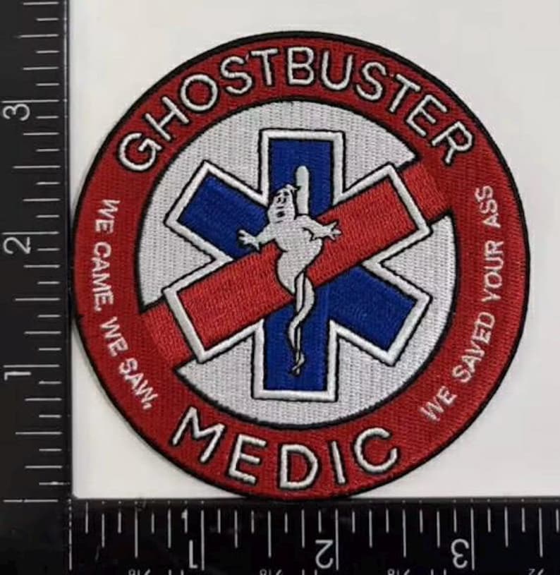K&ouml;nnte beinhalten: Ein kreisf&ouml;rmiger gestickter Aufn&auml;her mit rotem Rand und dem Text GHOSTBUSTER MEDIC sowie WE CAME, WE SAW, WE SAVED YOUR ASS. In der Mitte befindet sich ein blaues Stern-des-Lebens-Symbol mit einer wei&szlig;en Geisterfigur. Ein Lineal zeigt 8,9 cm.