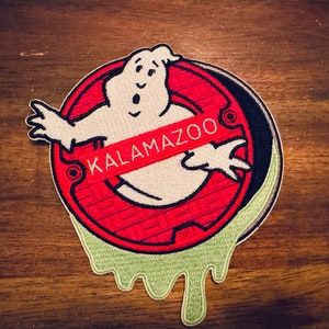 Puede incluir: Un parche bordado rojo y blanco con un fantasma con una pancarta roja "KALAMAZOO" sobre su cuerpo. El fantasma emerge de una rejilla de alcantarillado roja y negra con baba verde goteando de los bordes.