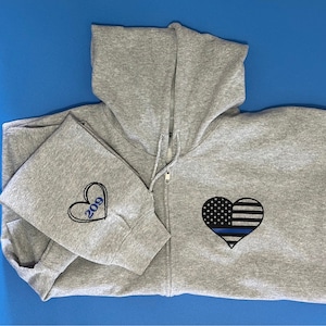 May include: A gray zip-up hoodie with a heart-shaped American flag design and a blue line. The left sleeve features a heart with the number "209". The hoodie has a hood and a zipper closure.