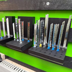 May include: A display of various reciprocating saw blades, arranged on black metal shelves against a green and black striped background. The blades are silver with visible teeth, some with colored bases. The display showcases different blade types for woodworking and metalworking.