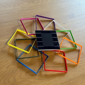 Könnte beinhalten: Eine Sammlung farbenfroher quadratischer Rahmen in verschiedenen Farben, darunter Rosa, Lila, Weiß, Grün, Orange, Gelb, Blau und Rot, die ein schwarzes rechteckiges Objekt mit einem Rastermuster umgeben. Die Artikel sind auf einer Holzoberfläche angeordnet.