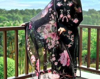 Op de afbeelding: Zwart salwar kameez-pak met bloemenprint en bijpassende dupatta. De outfit heeft roze en witte bloemenborduursels en een doorschijnende dupatta. Het model staat op een balkon met een groene achtergrond.