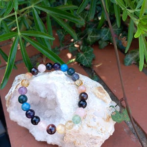 Puede incluir: Un brazalete de cuentas de colores con varias piedras preciosas, incluyendo amatista, cuarzo rosa, citrino y ónix negro. El brazalete se muestra sobre una roca blanca con una formación de cristal natural.