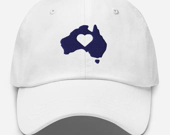 Op de afbeelding: Witte baseballpet met een marineblauw geborduurd silhouet van Australië met een hartvormige uitsparing in het midden. De pet heeft een gebogen klep en een verstelbare band aan de achterkant.