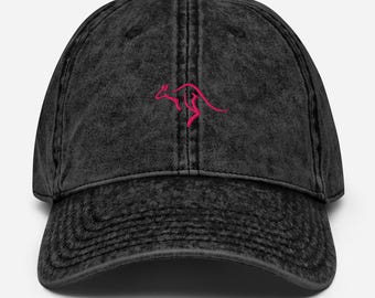 Op de afbeelding: Zwarte baseballcap in vintage-stijl met een gebogen klep. De cap heeft een roze geborduurd kangoeroe-logo op de voorkant. De cap heeft een distressed look, met een knoop bovenop en een verstelbare band aan de achterkant.