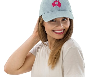 Op de afbeelding: Lichtblauwe denim baseballcap met een roze omtrek van Australië en een hart in het midden. De cap heeft een gebogen klep en een verstelbare band. De hoed wordt door een persoon gedragen.