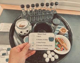 May include: A Halloween-themed science experiment set with a black tray, a glass jar with a jack-o'-lantern face, a small bowl of colorful beads, a small bowl of white cotton balls, and a card that reads "Moon Dust" with instructions for a science experiment. The instructions include 1 moon, 5 shakes of glitter, 2 soap pumps, 5 scoops of baking soda, and 3 pours of vinegar.