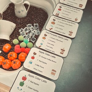 K&ouml;nnte beinhalten: Ein sensorisches Spielset mit einem wei&szlig;en Milchk&auml;nnchen, einer wei&szlig;en Tasse, dunkelbraunen Kaffeebohnen, durchsichtigen Acryl-Eisw&uuml;rfeln und kleinen orangefarbenen K&uuml;rbisfiguren. Sechs Rezeptkarten listen Zutaten wie Pumpkin Spice Latte und Apple Cider Latte mit genauen Mengenangaben f&uuml;r L&ouml;ffel, &Auml;pfel, Eisw&uuml;rfel und Zimtstangen auf.