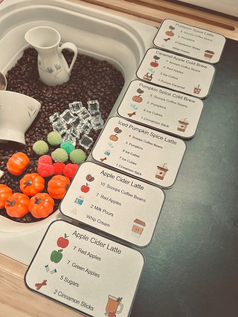 K&ouml;nnte beinhalten: Ein sensorisches Spielset mit einem wei&szlig;en Milchk&auml;nnchen, einer wei&szlig;en Tasse, dunkelbraunen Kaffeebohnen, durchsichtigen Acryl-Eisw&uuml;rfeln und kleinen orangefarbenen K&uuml;rbisfiguren. Sechs Rezeptkarten listen Zutaten wie Pumpkin Spice Latte und Apple Cider Latte mit genauen Mengenangaben f&uuml;r L&ouml;ffel, &Auml;pfel, Eisw&uuml;rfel und Zimtstangen auf.