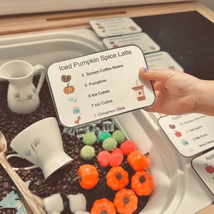 May include: A rectangular recipe card titled Iced Pumpkin Spice Latte with a list of ingredients including coffee beans, pumpkins, ice cubes, and a cinnamon stick, accompanied by a white ceramic pitcher, a teacup, and various small sensory play items.