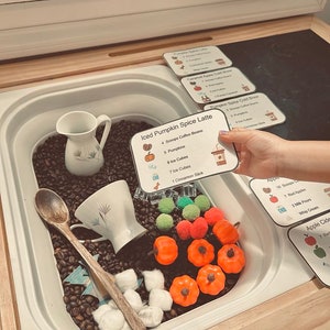 K&ouml;nnte beinhalten: Eine Sensorik-Box gef&uuml;llt mit dunklen Kaffeebohnen, kleinen orangefarbenen Kunststoffk&uuml;rbissen, gr&uuml;nen und orangefarbenen Pompons, wei&szlig;en Watteb&auml;llchen, einem Holzl&ouml;ffel, einem wei&szlig;en Keramikkrug und einer wei&szlig;en Teetasse mit Rezeptkarten.