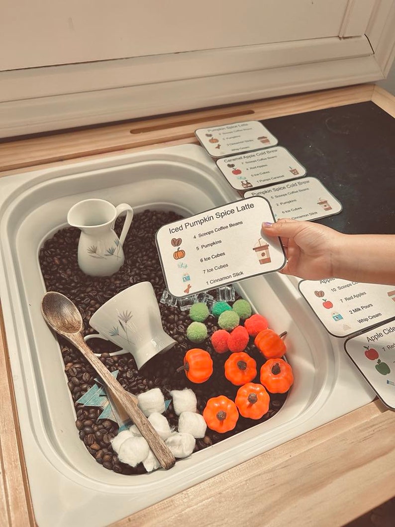 K&ouml;nnte beinhalten: Eine Sensorik-Box gef&uuml;llt mit dunklen Kaffeebohnen, kleinen orangefarbenen Kunststoffk&uuml;rbissen, gr&uuml;nen und orangefarbenen Pompons, wei&szlig;en Watteb&auml;llchen, einem Holzl&ouml;ffel, einem wei&szlig;en Keramikkrug und einer wei&szlig;en Teetasse mit Rezeptkarten.