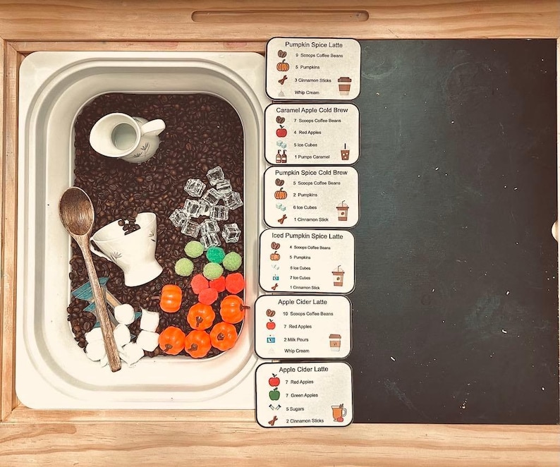 K&ouml;nnte beinhalten: Eine Sensorik-Box gef&uuml;llt mit dunklen Kaffeebohnen, einem Holzl&ouml;ffel, einem wei&szlig;en Keramikkrug, einer wei&szlig;en Teetasse, durchsichtigen Acryl-Eisw&uuml;rfeln, kleinen orangefarbenen K&uuml;rbissen und gr&uuml;nen Pompons. Sechs Rezeptkarten listen Zutaten f&uuml;r Getr&auml;nke auf.