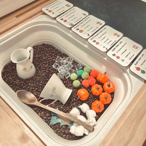 K&ouml;nnte beinhalten: Eine sensorische Spielkiste gef&uuml;llt mit Kaffeebohnen, einem wei&szlig;en Keramikkrug, einer wei&szlig;en Teetasse, einem Holzl&ouml;ffel, durchsichtigen Acryl-Eisw&uuml;rfeln, kleinen orangefarbenen Kunststoffk&uuml;rbissen, gr&uuml;nen und orangefarbenen Pompons und wei&szlig;en Watteb&auml;llchen.