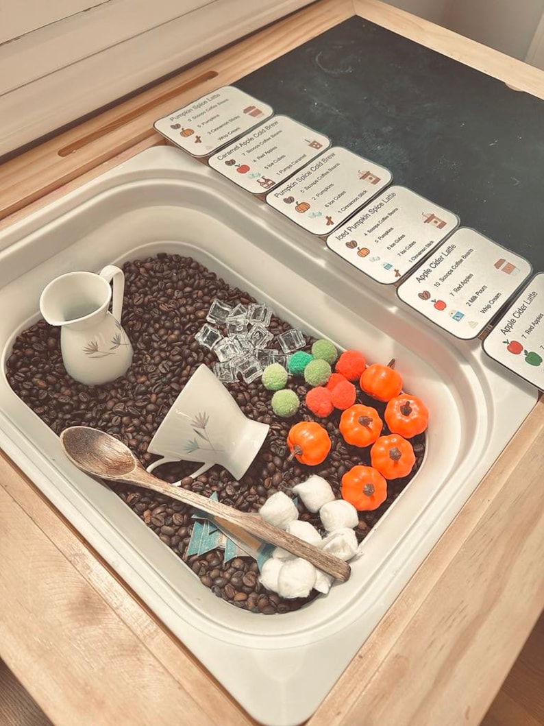 K&ouml;nnte beinhalten: Eine sensorische Spielkiste gef&uuml;llt mit Kaffeebohnen, einem wei&szlig;en Keramikkrug, einer wei&szlig;en Teetasse, einem Holzl&ouml;ffel, durchsichtigen Acryl-Eisw&uuml;rfeln, kleinen orangefarbenen Kunststoffk&uuml;rbissen, gr&uuml;nen und orangefarbenen Pompons und wei&szlig;en Watteb&auml;llchen.