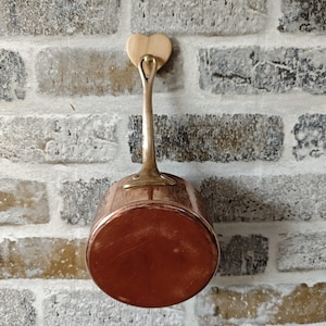 Könnte beinhalten: Ein Kupfertopf mit Messinggriff und einem herzförmigen Holzhalter. Der Topf hängt an einer Backsteinmauer. Der Topf hat einen Durchmesser von etwa 15 cm.