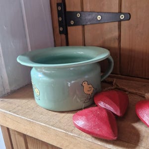 May include: A light green enamel chamber pot featuring a small yellow chick illustration on the side. The pot has a wide rim and a single handle. Two red wooden heart-shaped ornaments are positioned in the foreground near the base of the pot.