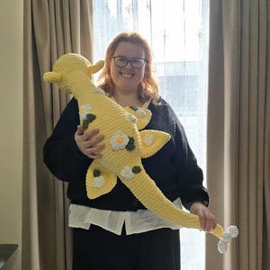 May include: A large, yellow crocheted sea serpent plush toy with white and green floral accents. The toy has a long, curved neck and tail, and is being held up by a person. The plush is approximately 40 inches long.
