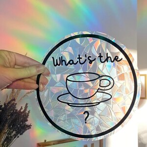 May include: A round, iridescent window cling with a black border and the text "What's the" above a teacup and saucer graphic. A question mark is below the teacup. The cling is held up in front of a rainbow light.