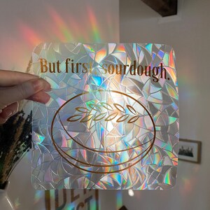May include: Iridescent square window cling with the text "But first, sourdough." A gold illustration of a loaf of bread with wheat is below the text. The cling is held up by a hand, with light refracting through it.