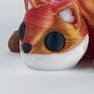 May include: A small, 3D printed fox figurine with a curled body and a white face. The fox is orange and red with black eyes and a white nose.