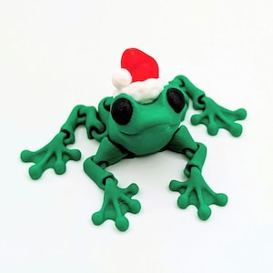 May include: A green articulated frog figurine wearing a red Santa hat with a white pom-pom. The frog has large black eyes and is posed on a white surface. The frog's limbs are jointed.