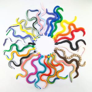 May include: A collection of colorful, flexible snake toys arranged in a circular pattern. The snakes come in various colors, including blue, green, yellow, red, and pink, with some featuring patterned designs. The toys are made of a soft, bendable material.