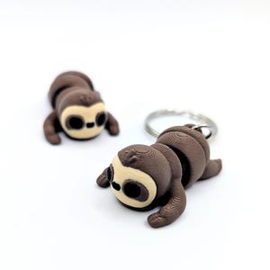 May include: Two brown sloth keychains with a light tan face and black eyes are displayed on a white background. One keychain is attached to a silver ring. The sloths are lying down, and the details of their bodies are visible.